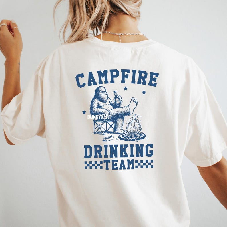 Puede incluir: Camiseta color crema con un gr&aacute;fico azul de una criatura parecida a Bigfoot relaj&aacute;ndose junto a una fogata, sosteniendo una botella. El texto "CAMPFIRE DRINKING TEAM" est&aacute; arriba y abajo de la ilustraci&oacute;n. La camiseta es de corte casual.