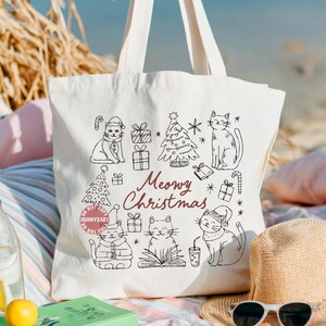 May include: Off-white canvas tote bag featuring a black and white illustration of cats, Christmas trees, and gifts. The words "Meowy Christmas" are written in a burgundy script. The bag has two white handles.