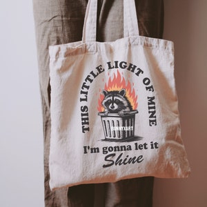 May include: Cream-colored tote bag featuring a raccoon in a trash can surrounded by flames. The bag has the text "THIS LITTLE LIGHT OF MINE" arched above the raccoon, and "I'm gonna let it Shine" below. The brand name "BunnyXart" is at the bottom.