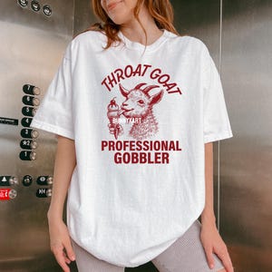 May include: White t-shirt with a red graphic of a goat eating ice cream. The text on the shirt reads "Throat Goat Professional Gobbler".