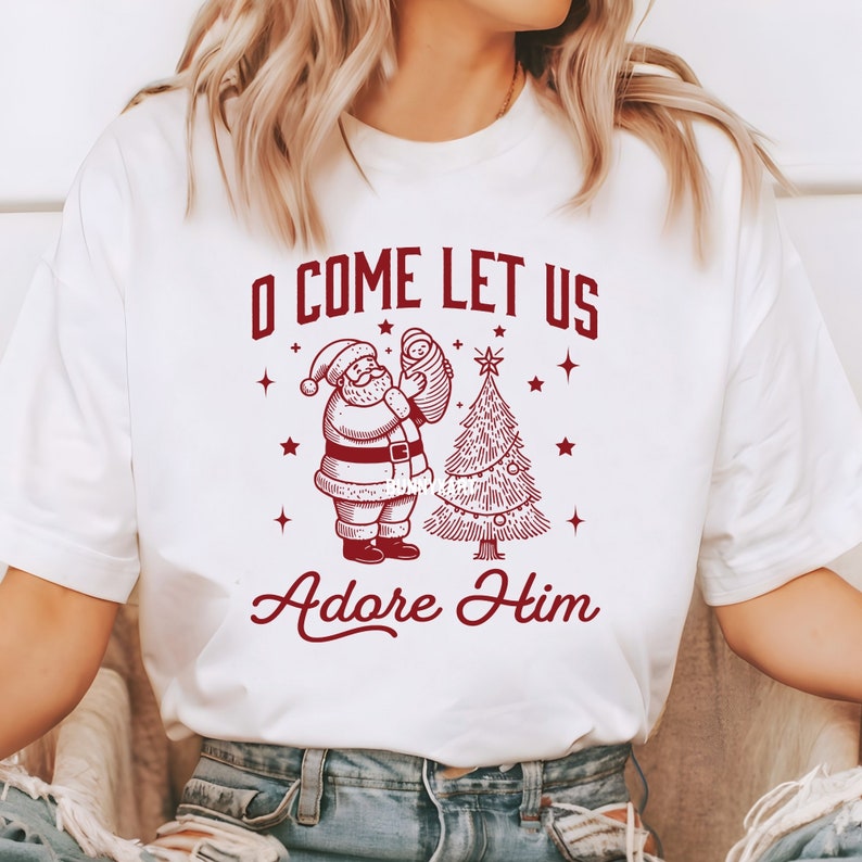 Pode incluir: Camiseta branca com um gr&aacute;fico vermelho apresentando o Papai Noel segurando um beb&ecirc; ao lado de uma &aacute;rvore de Natal. O texto "O COME LET US Adore Him" est&aacute; acima e abaixo da imagem. O design inclui estrelas.