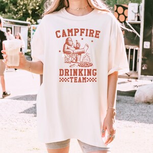 Puede incluir: Camiseta color crema con un gr&aacute;fico rojo que dice "Campfire Drinking Team". El gr&aacute;fico presenta un personaje de Bigfoot relaj&aacute;ndose junto a una fogata, sosteniendo una cerveza. La camiseta tiene un corte holgado.