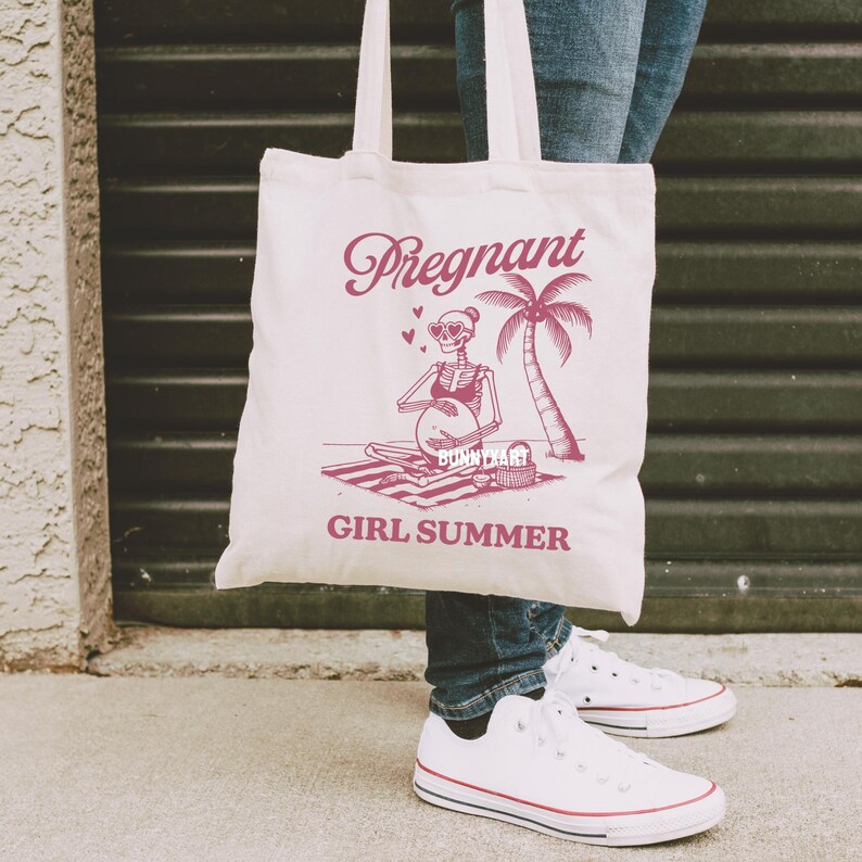 May include: A white tote bag featuring a pink graphic of a skeleton wearing sunglasses and holding a beach ball. The text "Pregnant Girl Summer" is printed on the bag.