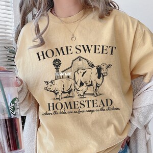Pode incluir: Uma camiseta amarela com um design gr&aacute;fico preto que diz "Home Sweet Homestead where the kids are as free range as the chickens". O gr&aacute;fico tamb&eacute;m inclui uma casa de fazenda, uma vaca, um porco e uma galinha.