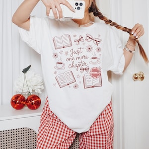 Op de afbeelding: Wit T-shirt met een bordeauxrode opdruk met de tekst "Just one more chapter". Het ontwerp bevat open boeken, koffiekopjes, strikken en brillen. De persoon draagt een rood-witte geruite broek.