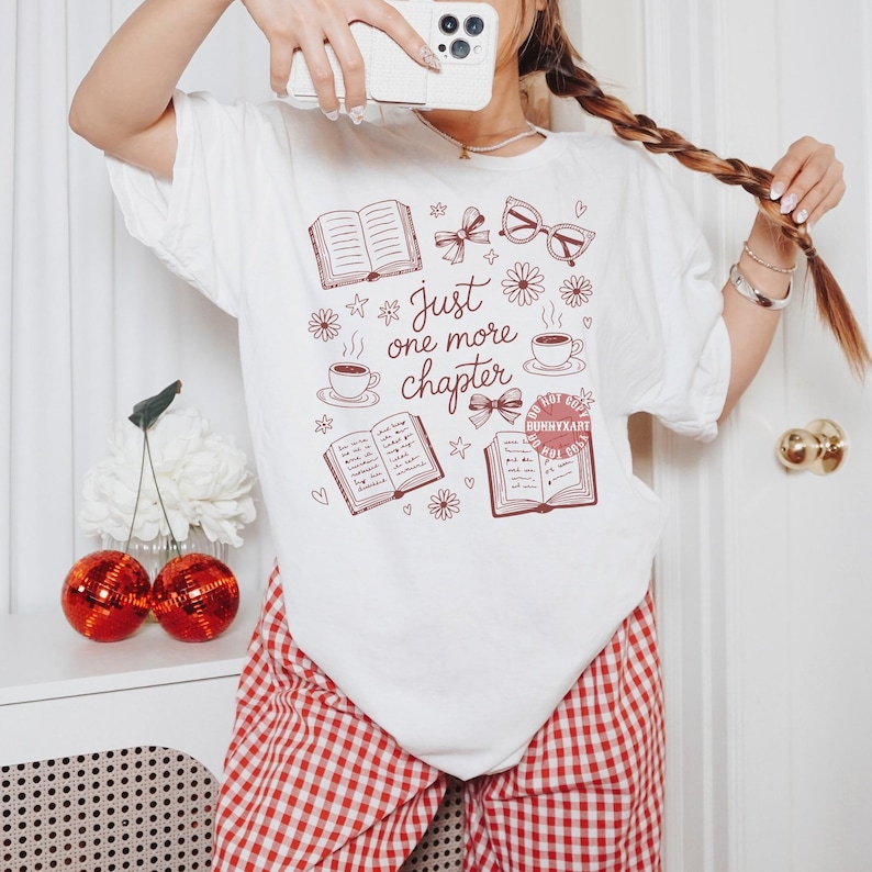 Op de afbeelding: Wit T-shirt met een bordeauxrode opdruk met de tekst "Just one more chapter". Het ontwerp bevat open boeken, koffiekopjes, strikken en brillen. De persoon draagt een rood-witte geruite broek.