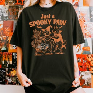May include: Black t-shirt with an orange graphic of three dogs dressed in Halloween costumes. The text "Just a SPOOKY PAW" is above the dogs. The brand name "BUNNYXART PRINTS" is below the dogs.