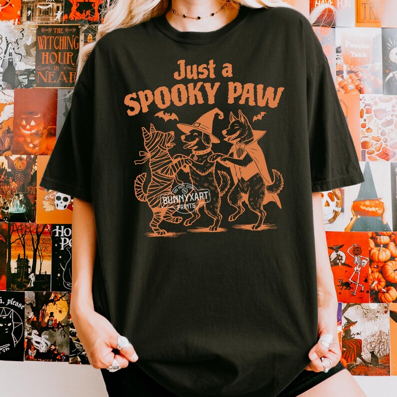May include: Black t-shirt with an orange graphic of three dogs dressed in Halloween costumes. The text "Just a SPOOKY PAW" is above the dogs. The brand name "BUNNYXART PRINTS" is below the dogs.