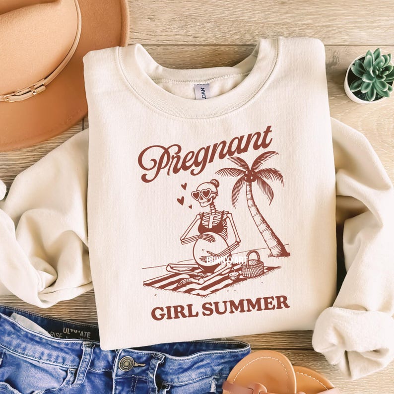 May include: A beige sweatshirt with a red graphic of a skeleton wearing sunglasses and a bikini, holding a beach ball. The text "Pregnant Girl Summer" is printed below the graphic.