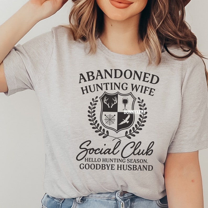 Op de afbeelding: Grijs T-shirt met zwarte tekst die "Abandoned Hunting Wife Social Club Hello Hunting Season Goodbye Husband" luidt. De tekst wordt omringd door een zwart-wit wapenschild met jachtgerelateerde afbeeldingen.