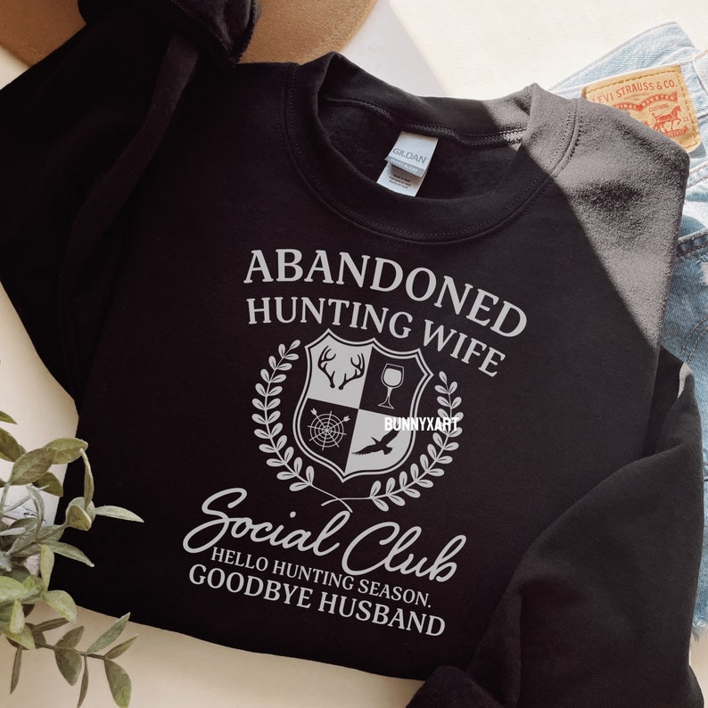 Op de afbeelding: Zwart sweatshirt met witte tekst die "Abandoned Hunting Wife Social Club Hello Hunting Season. Goodbye Husband" luidt. De tekst wordt omringd door een wit wapenschild met een hert, een wijnglas, een kompas en een vogel.