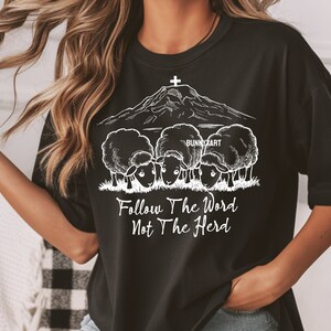 May include: Black t-shirt with a white graphic of a mountain with a cross on top and three sheep in front of it. The text "Follow The Word Not The Herd" is below the sheep.