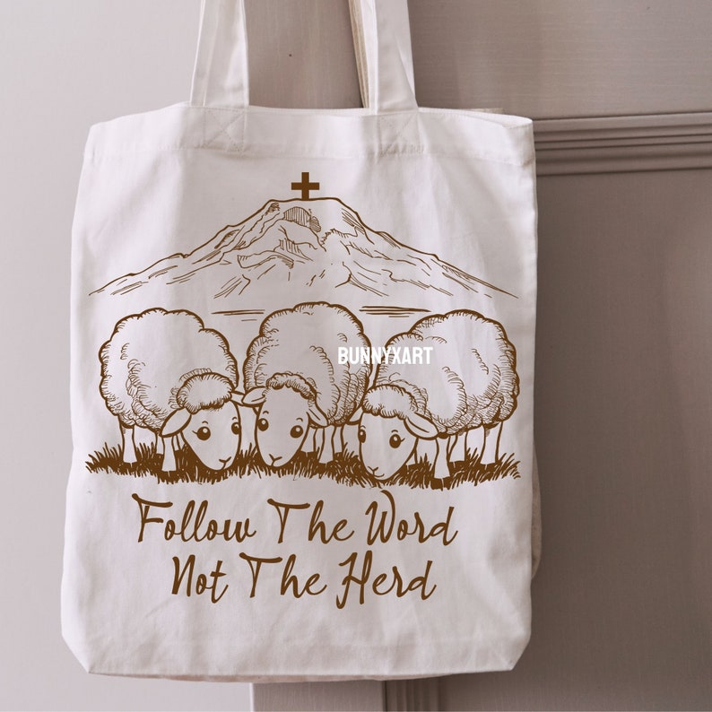 May include: A white tote bag with a brown illustration of a mountain with a cross on top and four sheep grazing at the bottom. The text "Follow The Word Not The Herd" is printed in brown below the sheep.