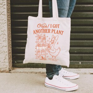 May include: A white canvas tote bag with the text "Oops! I Got Another Plant" in orange. The bag features an illustration of a duck holding a potted plant, with a plant shelf in the background. The bag has two white handles.