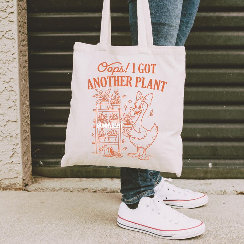 May include: A white canvas tote bag with the text "Oops! I Got Another Plant" in orange. The bag features an illustration of a duck holding a potted plant, with a plant shelf in the background. The bag has two white handles.