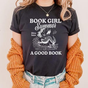 K&ouml;nnte beinhalten: Schwarzes T-Shirt mit dem Text "Book Girl Summer" und einer Grafik einer Meerjungfrau, die ein Buch liest. Der Text unter der Grafik lautet "Dive Into A Good Book".