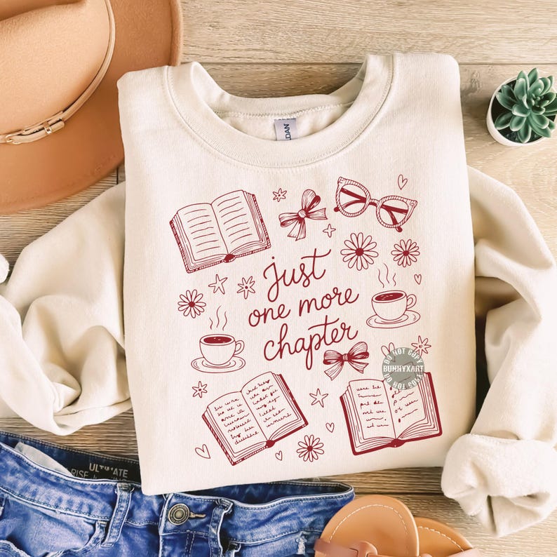 Op de afbeelding: Cr&egrave;mekleurige sweatshirt met een bordeauxrood grafisch ontwerp met boeken, koffiekopjes, strikken, brillen en bloemen. De tekst "Just one more chapter" is prominent aanwezig. De sweatshirt ligt op een houten oppervlak.