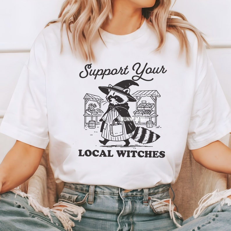 K&ouml;nnte beinhalten: Wei&szlig;es T-Shirt mit einer schwarz-wei&szlig;en Illustration eines Waschb&auml;ren, der einen Hexenhut tr&auml;gt und eine Einkaufstasche h&auml;lt. Der Text "Support Your Local Witches" ist unter der Illustration gedruckt.