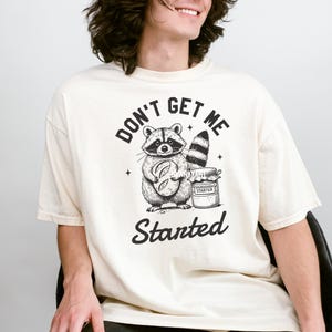 May include: A cream-colored t-shirt with a black graphic of a raccoon holding a loaf of bread and a jar of sourdough starter. The text "Don't Get Me Started" is printed in black below the graphic.