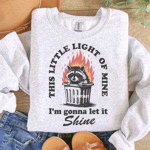 May include: Light gray sweatshirt with a graphic of a raccoon in a trash can surrounded by flames. The text reads "THIS LITTLE LIGHT OF MINE, I'M GONNA LET IT SHINE."