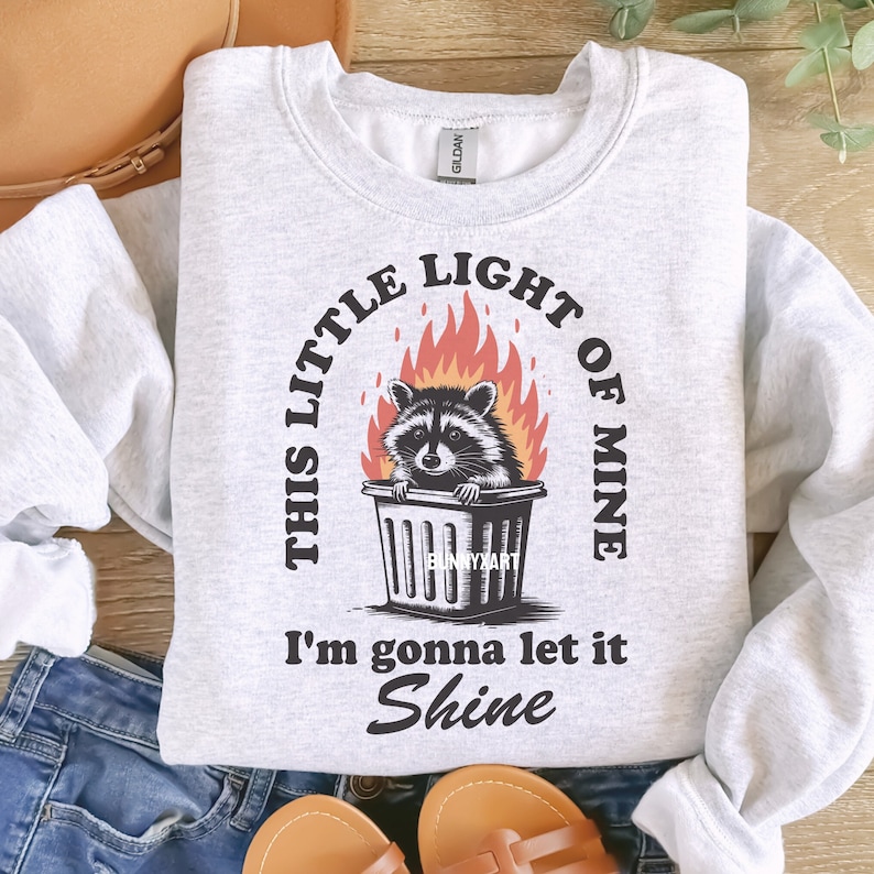 May include: Light gray sweatshirt with a graphic of a raccoon in a trash can surrounded by flames. The text reads "THIS LITTLE LIGHT OF MINE, I'M GONNA LET IT SHINE."