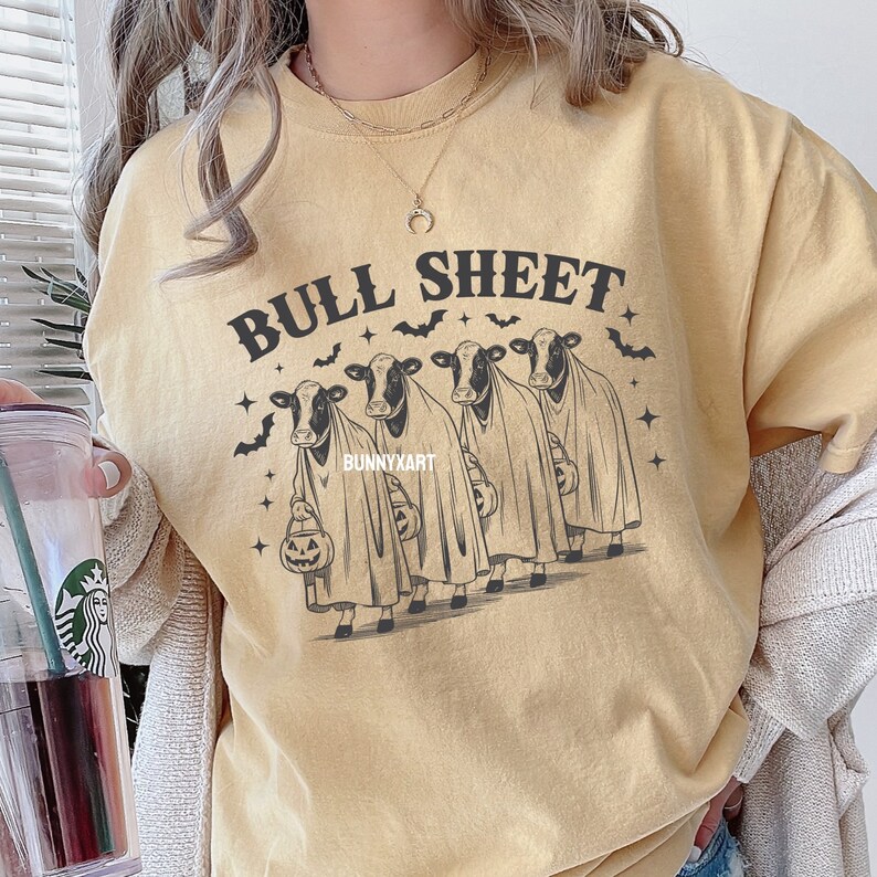 May include: Mustard yellow t-shirt with the phrase "BULL SHEET" in dark letters. The graphic features four cows dressed as ghosts, holding jack-o'-lantern buckets, with bats and stars. The artist's name, "BUNNYXART", is also visible.