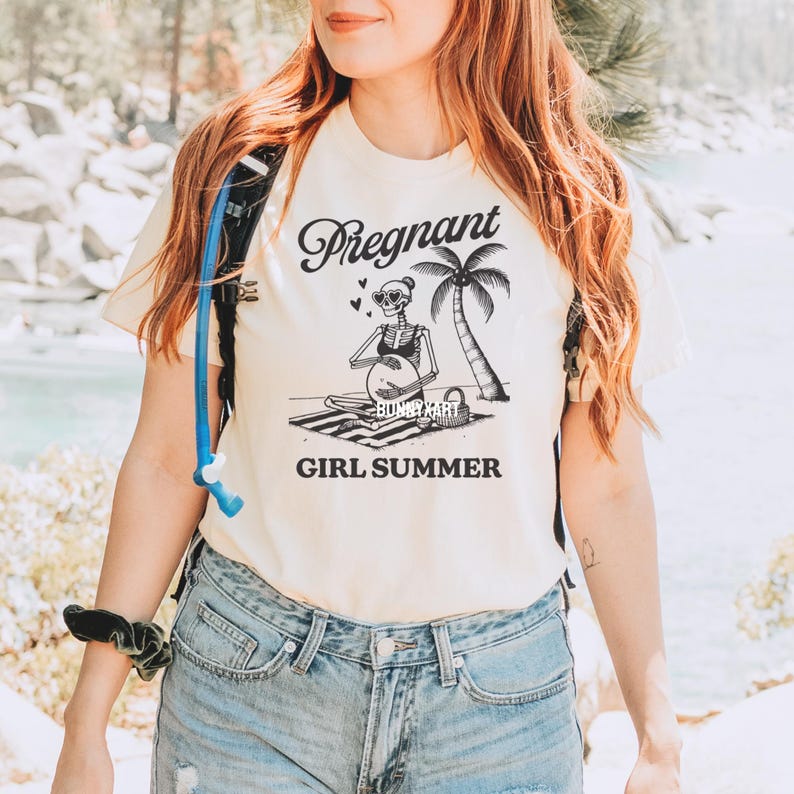 May include: A white t-shirt featuring a black graphic of a skeleton wearing sunglasses and a bikini, holding a beach ball. The text "Pregnant Girl Summer" is printed below the graphic.