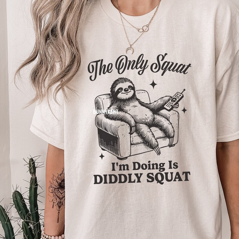 May include: A white t-shirt featuring a black graphic of a sloth relaxing in an armchair with a remote control. The text "The Only Squat I'm Doing Is Diddly Squat" is printed beneath the sloth.