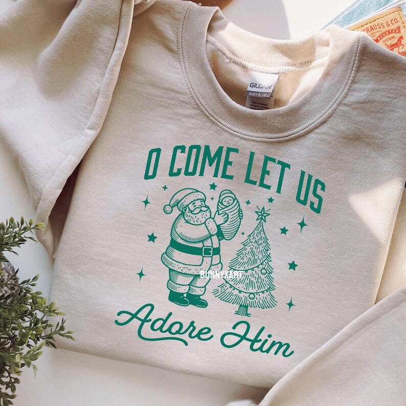 Pode incluir: Camisola bege de gola redonda com um gr&aacute;fico verde. O gr&aacute;fico apresenta o Pai Natal segurando um beb&eacute;, uma &aacute;rvore de Natal e o texto "O COME LET US Adore Him". O design inclui estrelas e elementos decorativos.