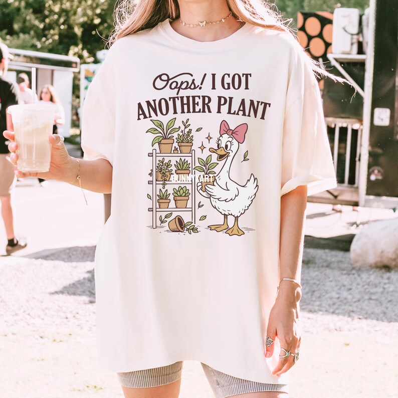 May include: Cream-colored t-shirt with the text "Oops! I Got Another Plant." The graphic features a cartoon duck with a pink bow, holding a plant, and a shelf with potted plants. The design includes the text "Bunny Art."