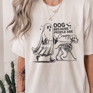 May include: Cream-coloured t-shirt with a black and white graphic of a ghost walking a skeleton dog on a lead. The text reads "DOG BECAUSE PEOPLE ARE Creepy" with bat and star accents. The shirt has a crew neck.