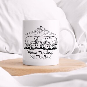 May include: White ceramic mug with a black and white illustration of a mountain with a cross on top and four sheep looking down at the text "Follow The Word Not The Herd".