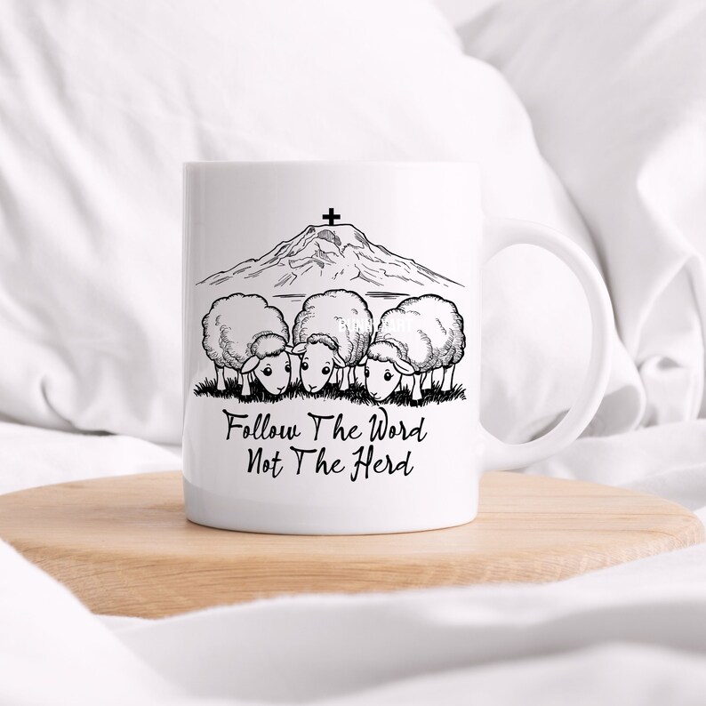 May include: White ceramic mug with a black and white illustration of a mountain with a cross on top and four sheep looking down at the text "Follow The Word Not The Herd".