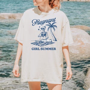 May include: A white t-shirt featuring a blue graphic of a skeleton wearing sunglasses and a bikini, sitting on a beach towel with a palm tree and the text "Pregnant Girl Summer".
