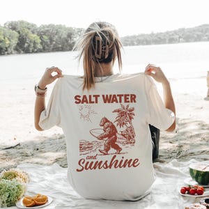 May include: Cream-colored t-shirt with a graphic of a Bigfoot carrying a surfboard, palm trees, and the words "Salt Water and Sunshine" in a reddish-brown color. The shirt is being held up by a person.