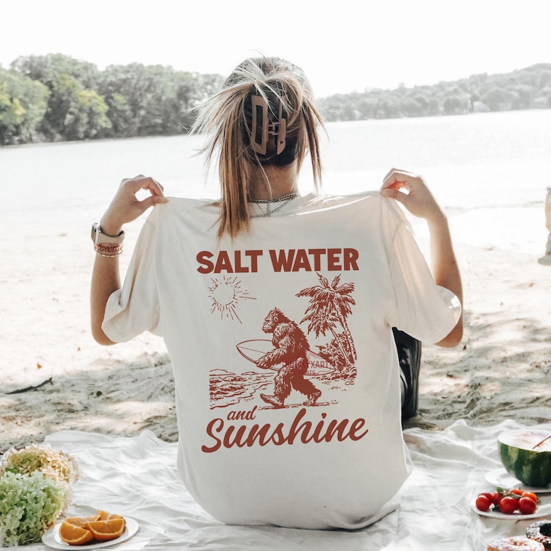 May include: Cream-colored t-shirt with a graphic of a Bigfoot carrying a surfboard, palm trees, and the words "Salt Water and Sunshine" in a reddish-brown color. The shirt is being held up by a person.