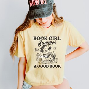 K&ouml;nnte beinhalten: Gelbes T-Shirt mit einem Grafikdruck einer Meerjungfrau, die ein Buch im Meer liest. Der Text auf dem T-Shirt lautet "Book Girl Summer Dive Into A Good Book".