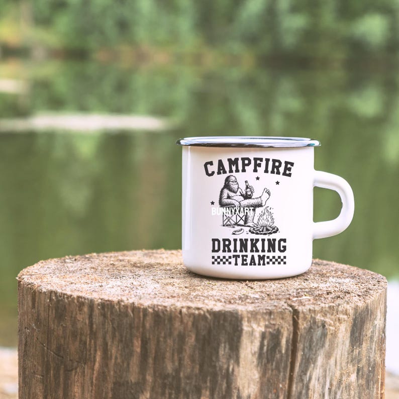 Puede incluir: Taza de camping de esmalte blanco con borde y asa negros. La taza presenta el texto "CAMPFIRE DRINKING TEAM" con un gr&aacute;fico de un personaje de Bigfoot relaj&aacute;ndose junto a una fogata. La taza est&aacute; sobre un toc&oacute;n de madera.