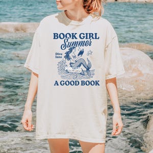 K&ouml;nnte beinhalten: Ein wei&szlig;es T-Shirt mit einer blauen Grafik einer Meerjungfrau, die auf einer Welle ein Buch liest. Der Text auf dem Shirt lautet "Book Girl Summer Dive Into A Good Book".