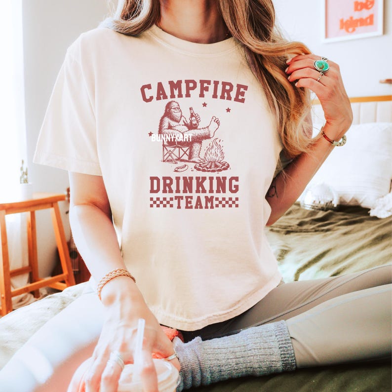 Puede incluir: Camiseta color crema con un gr&aacute;fico granate que dice "Campfire Drinking Team". El gr&aacute;fico presenta un personaje de Bigfoot relaj&aacute;ndose junto a una fogata, sosteniendo una cerveza. La camiseta es de estilo casual y de manga corta.