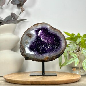 May include: A polished amethyst geode slice featuring deep purple crystalline structures at the center, surrounded by a natural grey and brown banded agate border. The geode is mounted on a black metal stand with a rectangular base.