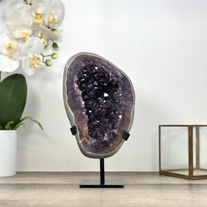 May include: A large amethyst geode, showcasing a deep purple crystal interior. The geode is mounted on a black metal stand, with a gray and brown outer edge. The geode is displayed on a wooden surface, with white orchids in the background.
