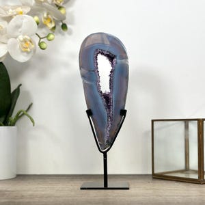 May include: A large, polished geode slice in shades of blue and purple, mounted on a black metal stand. The geode has a hollow centre filled with sparkling amethyst crystals. The overall shape is elongated and teardrop-like.