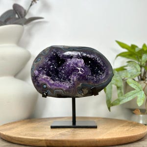 May include: A polished amethyst geode featuring a deep purple crystalline center surrounded by concentric bands of gray and black stone. The geode is mounted horizontally on a black metal stand with a square base, showcasing its natural formation.