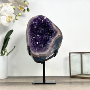 May include: A large amethyst geode, showcasing a deep purple crystal interior, is mounted on a black metal stand. The geode's outer edges display bands of gray and black. The stand has a square base and a vertical support.