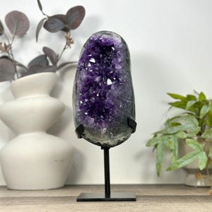 May include: A deep purple amethyst geode featuring jagged, crystalline structures within a natural stone cavity. The geode is securely mounted on a black metal stand with a vertical rod and a rectangular base, displaying its raw, textured exterior.