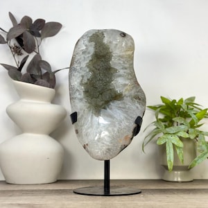 May include: A polished, irregular slab of translucent white quartz with a central, textured olive-green mineral inclusion. The stone is mounted vertically on a black metal stand with a circular base, showcasing its natural crystalline structure.