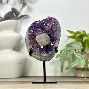 May include: A deep purple amethyst geode slice featuring a central translucent white calcite crystal formation. The geode is mounted on a thin black metal stand with a square base, displaying natural jagged edges and crystalline textures.
