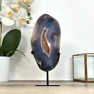 May include: A polished agate geode slice, presented on a black metal stand. The geode displays concentric bands of blue, grey, and brown tones, with a central cavity of crystalline formations. The stand is rectangular and has a single vertical support.