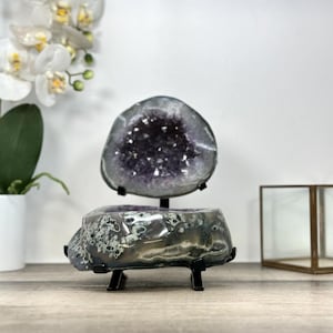 May include: A decorative geode with a dark purple crystal center, displayed on a black metal stand. The geode is a natural stone with a rough, textured exterior. A second geode is displayed above it. The background includes a white orchid and a glass display case.